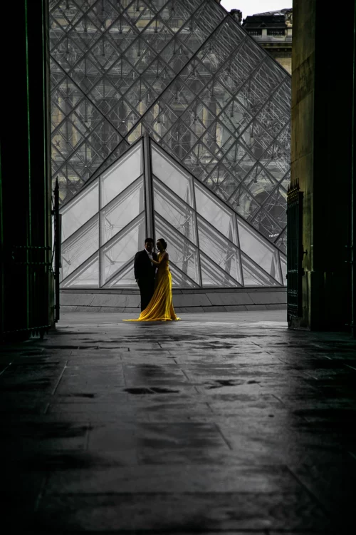 Alt Deux personnes, un homme en costume sombre et une femme en robe longue jaune, se tenant devant la pyramide du Louvre à Paris, vues à travers une arche sombre."