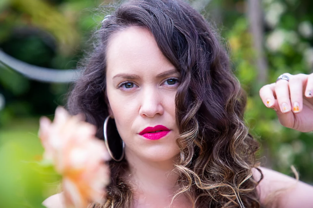 Alt Portrait rapproché d'une femme aux cheveux bouclés, boucles d'oreilles créoles, et rouge à lèvres rose vif, avec une fleur floue au premier plan et une main tendue sur le côté droit."