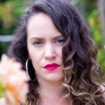 Alt Portrait rapproché d'une femme aux cheveux bouclés, boucles d'oreilles créoles, et rouge à lèvres rose vif, avec une fleur floue au premier plan et une main tendue sur le côté droit."